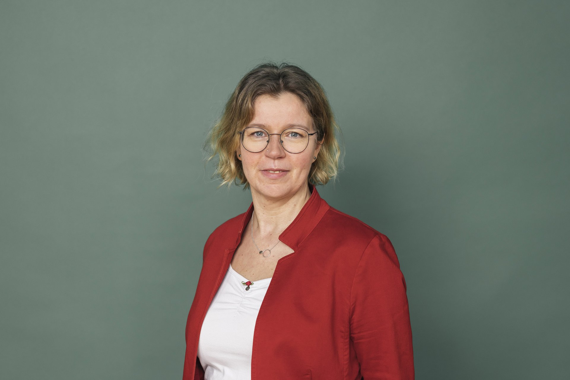Eine Frau mit Kinnlangen welligen blonden Haaren. Sie trägt eine Brille und lächelt. Sie trägt eine weißes Tshirt und eine rote Jacke darüber