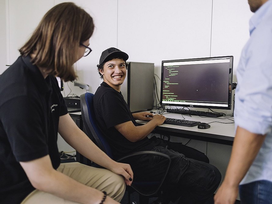 Ein junger Mann sitzt an einem Schreibtisch an einem Computer. Er dreht sich zu einem links neben ihm sitzenden Mitschüler und lacht ihn an. Sie befinden sich in einem Klassenraum