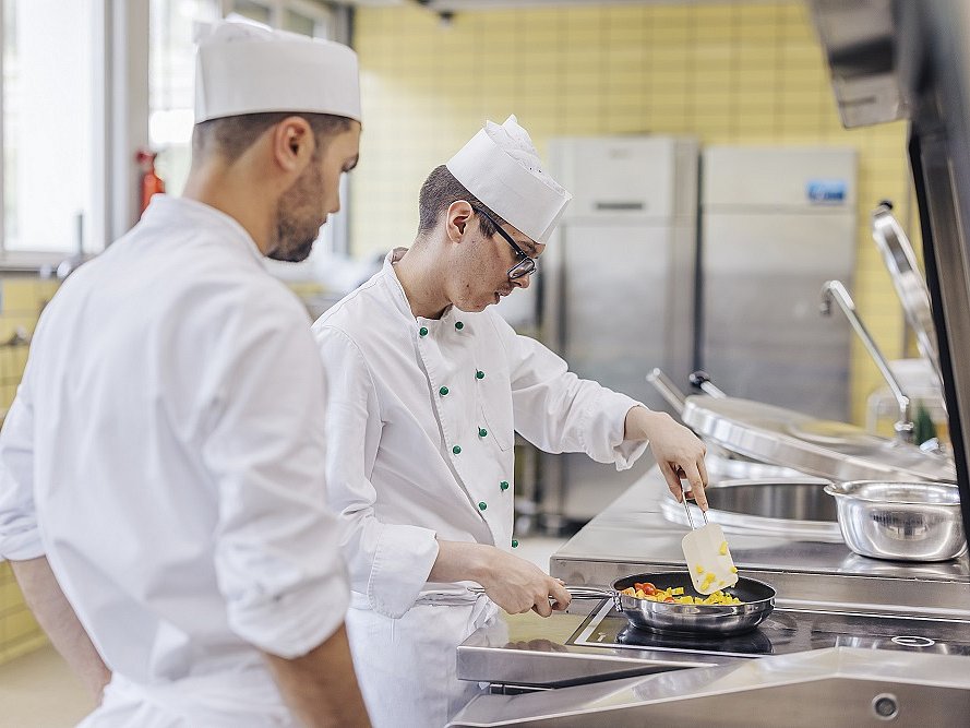 In einer Küche stehen Kochausbilder und Kochlehrling nebeneinander. Der Lehrling fertigt ein Gericht in einer Pfanne an. Beide tragen Arbeitskleidung und Kochmützen